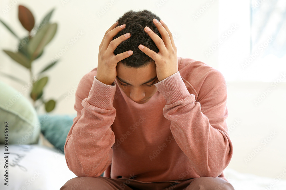 Sad African-American teenage boy sitting on bed at home