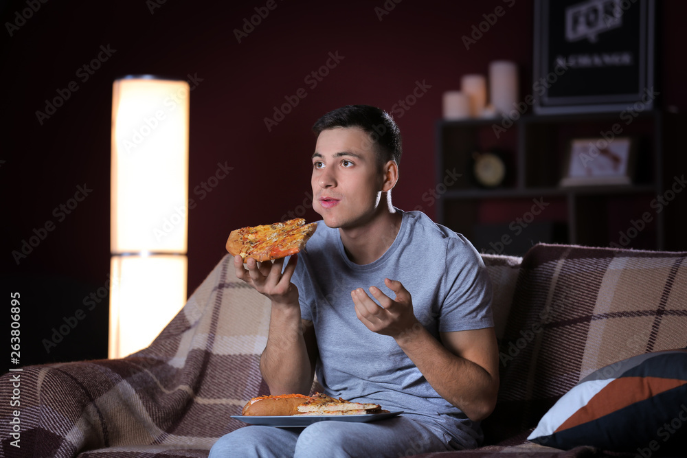 Handsome young man eating unhealthy food while watching TV at night