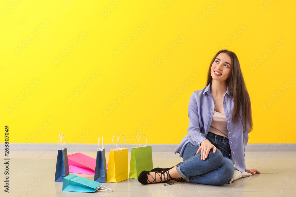 Thoughtful young woman with shopping bags sitting on floor near color wall