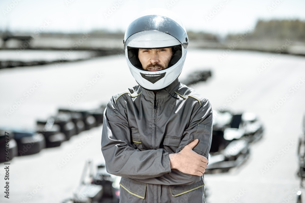 Portrait of a male racer in uniform and protective helmet standing on the track with karts on the ba