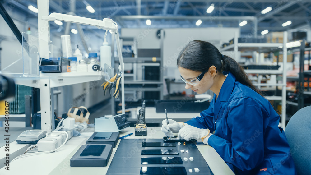 Young Female in Blue Work Coat is Assembling Printed Circuit Boards for Smartphones. Electronics Fac