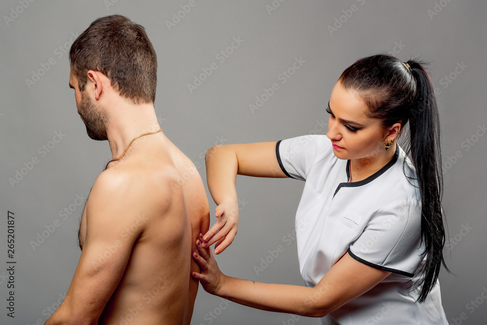 Beautiful nurse examining back of a man on the gray background. Brunette woman doctor in white medic