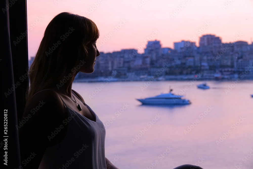 Woman in a white dress sitting on a window ledge, watching as the sun sets.