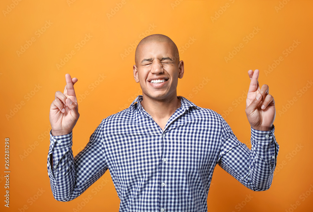 African-American man with crossed fingers making wish on color background