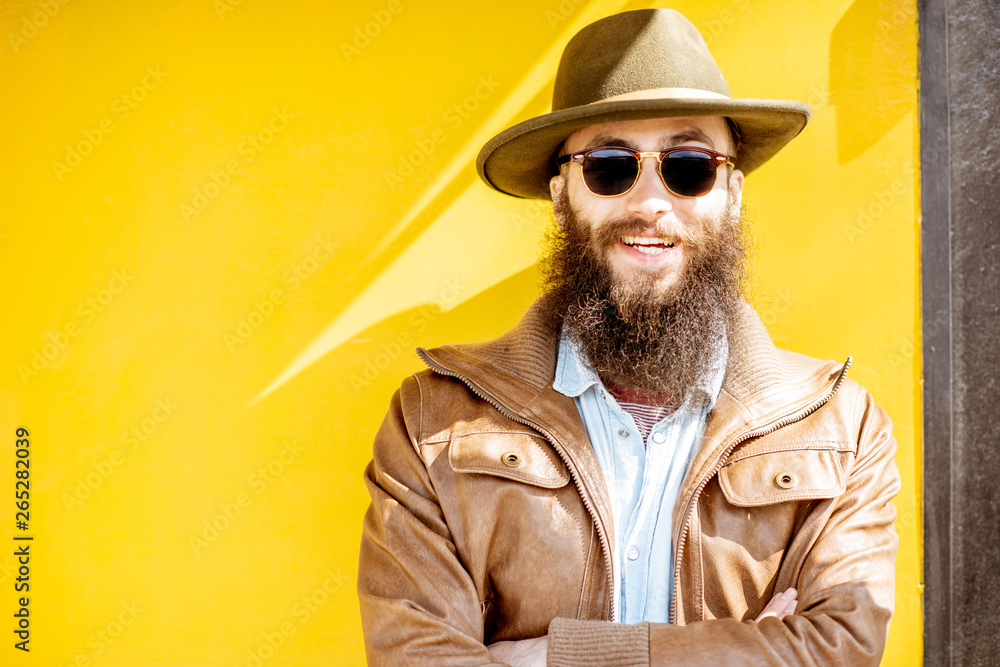 Portrait of a stylish bearded man dressed in jacket and hat on the bright yellow background outdoors