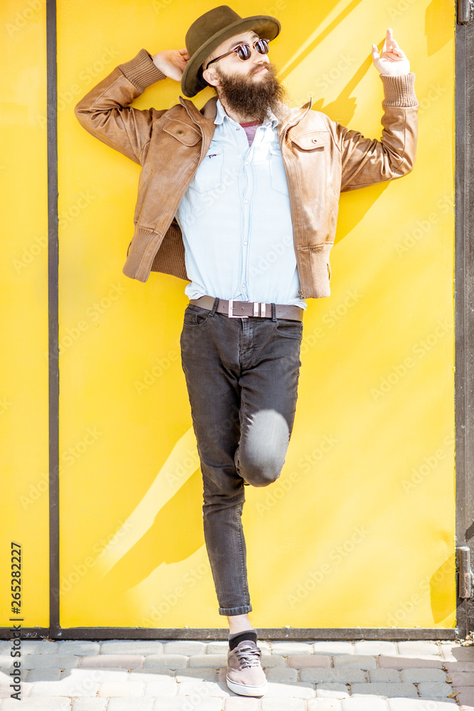 Full length portrait of a stylish bearded man dressed in jacket and hat on the bright yellow backgro