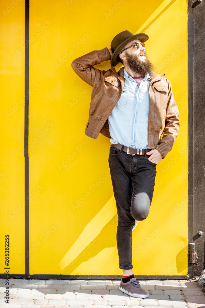 Full length portrait of a stylish bearded man dressed in jacket and hat on the bright yellow backgro