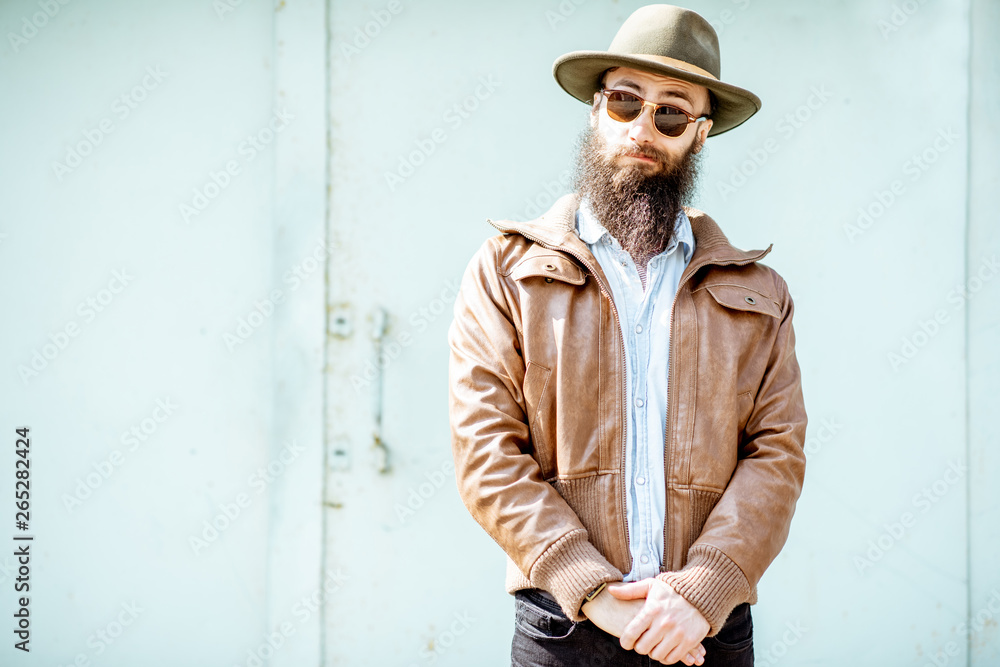 Portrait of a stylish bearded man dressed in jacket and hat on the light turquoise background outdoo