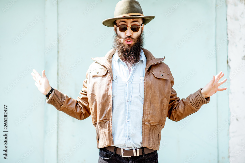 Portrait of a stylish bearded man dressed in jacket and hat on the light turquoise background outdoo