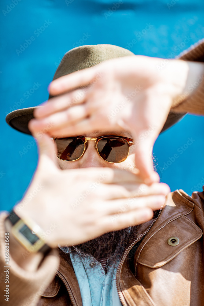 Close-up portrait of a stylish bearded man with hat and sunglasses on the blue wall background