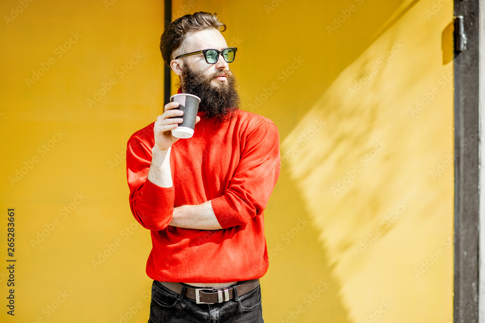 Portrait of a stylish bearded man in red swewater standing with coffee cup on the yellow background