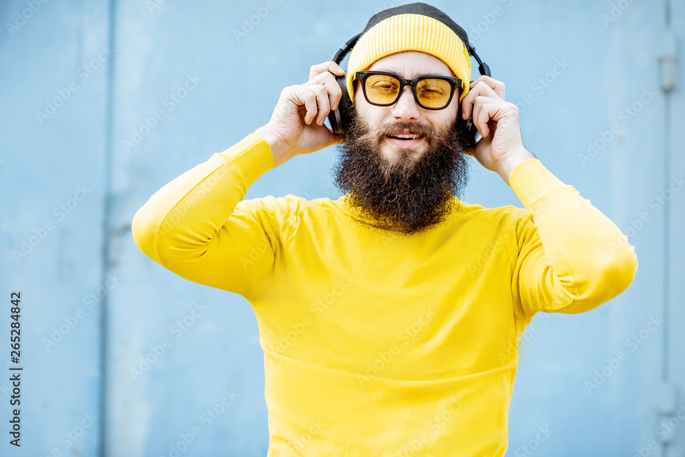 Portrait of a stylish bearded man in yellow clothes enjoying music with headphones on the blue backg