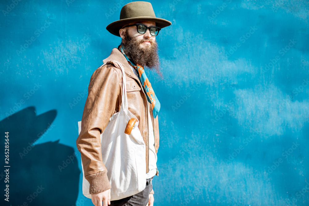 Portrait of a stylish bearded man dressed in jacket and hat standing with bag on the blue background