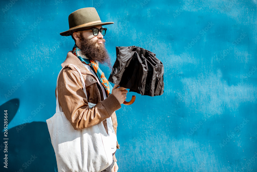 Portrait of a stylish bearded man dressed in jacket and hat standing with bag on the blue background