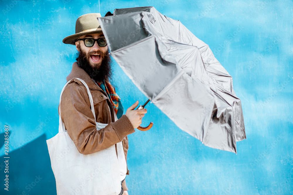 Portrait of a stylish bearded man dressed in jacket and hat standing with bag on the blue background