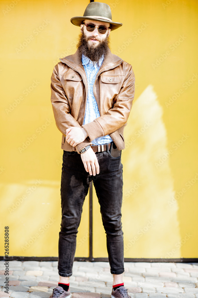 Full length portrait of a stylish bearded man dressed in jacket and hat on the bright yellow backgro