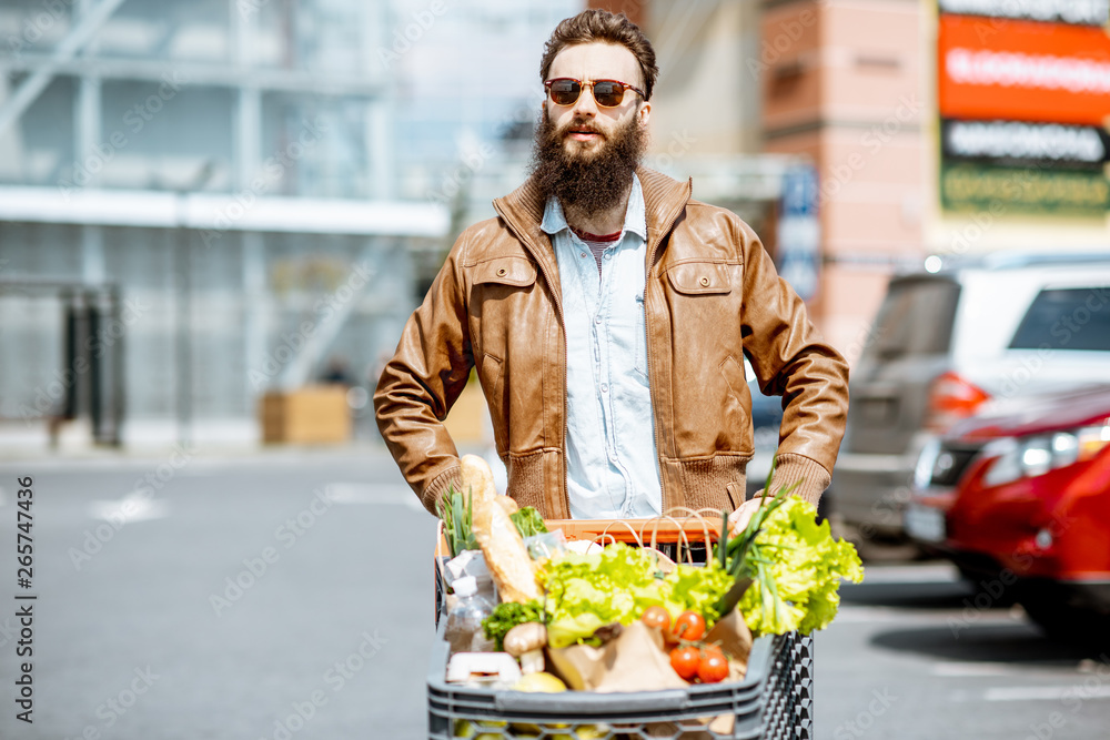 超市附近的户外停车场上，一个时尚的男人拿着装满新鲜健康食品的购物车