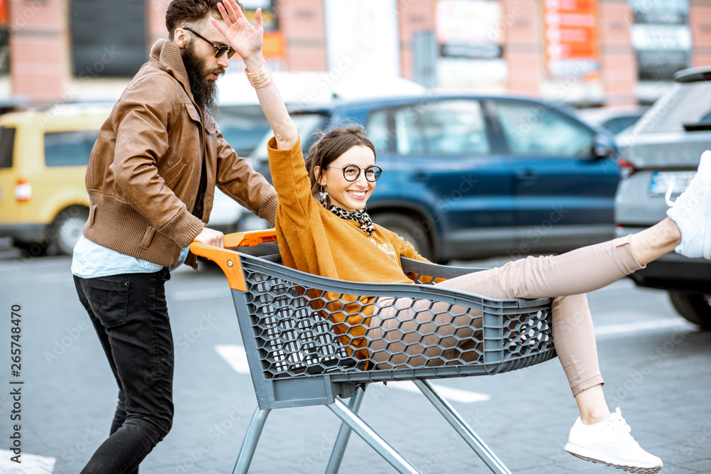 Young stylish coupe having fun riding with shopping cart on the outdoor parking near the supermarket