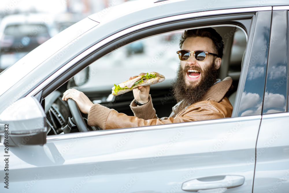 Stylish bearded man having a snack with tasty sandwich while driving a car in the city