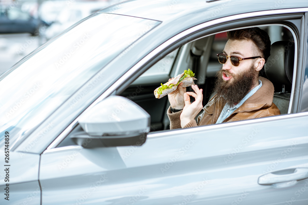 Stylish bearded man having a snack with tasty sandwich while driving a car in the city