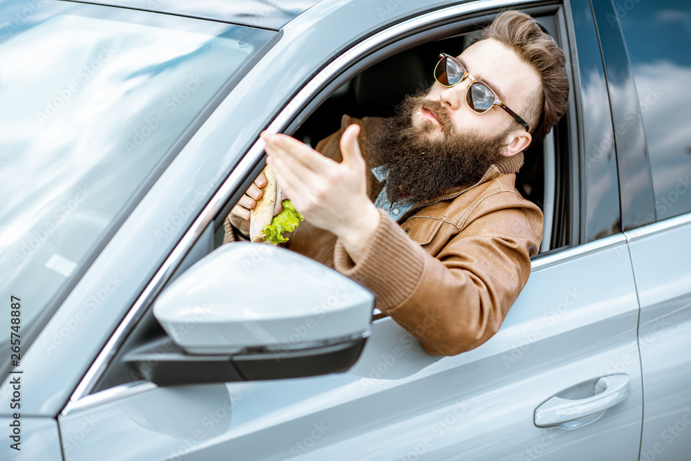 Stylish bearded man having a snack with tasty sandwich while driving a car in the city