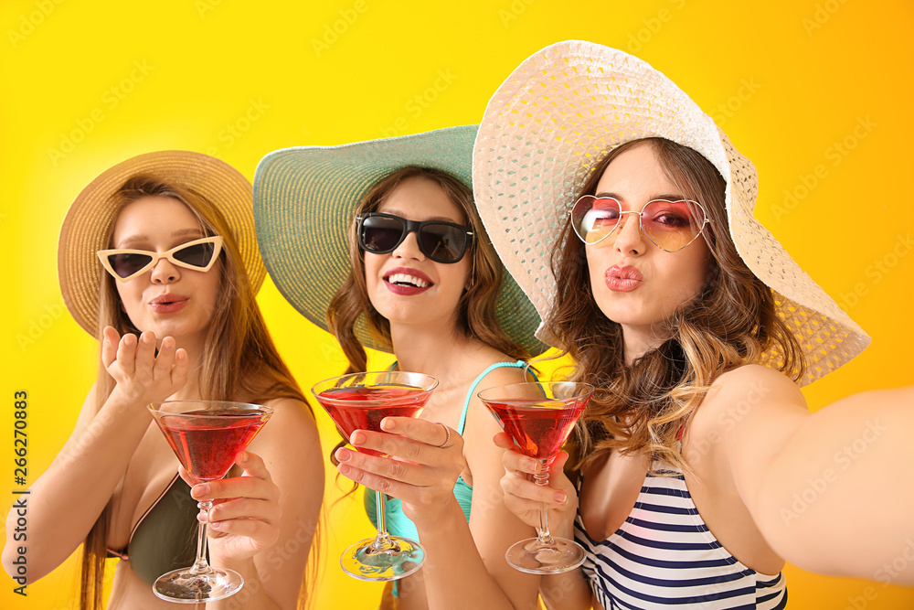 Beautiful young women in swimsuits taking selfie with cocktails on color background