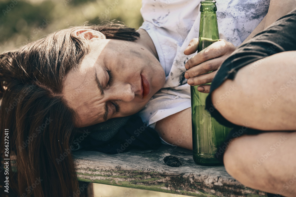 可怜的无家可归的女人拿着瓶子躺在户外的长椅上