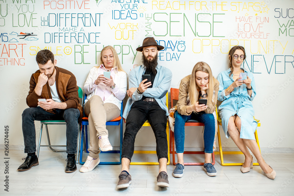 Group of diverse people using smart phones while sitting together in a row on the wall background in