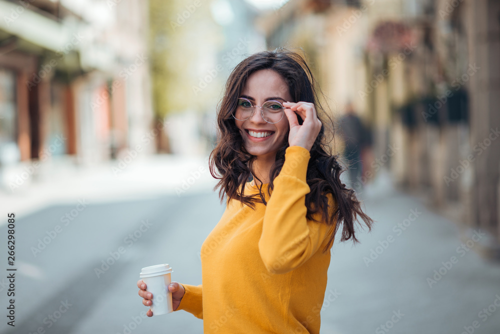 一个阳光明媚的日子里，带着外卖咖啡在城市里散步的美丽年轻女子。