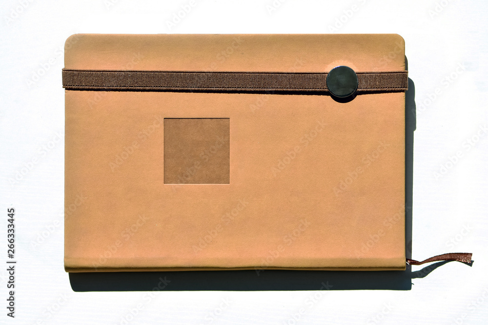 Notebook (beige, brown), lying on a white table brightly lit by natural daylight.