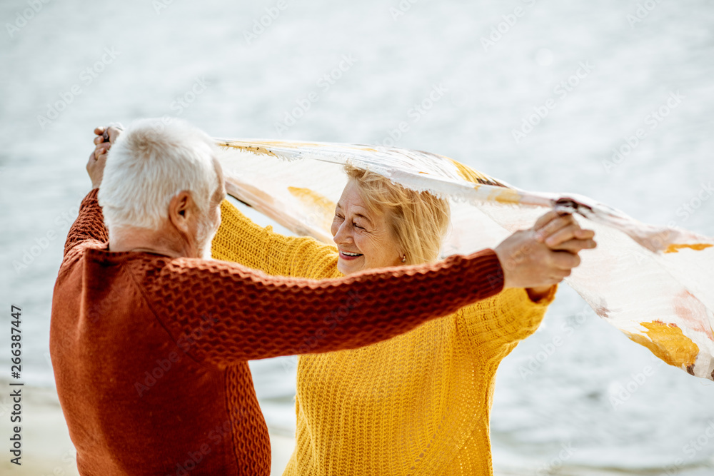 Lovely senior couple playing with scarf during the windy weather, standing together on the sandy bea