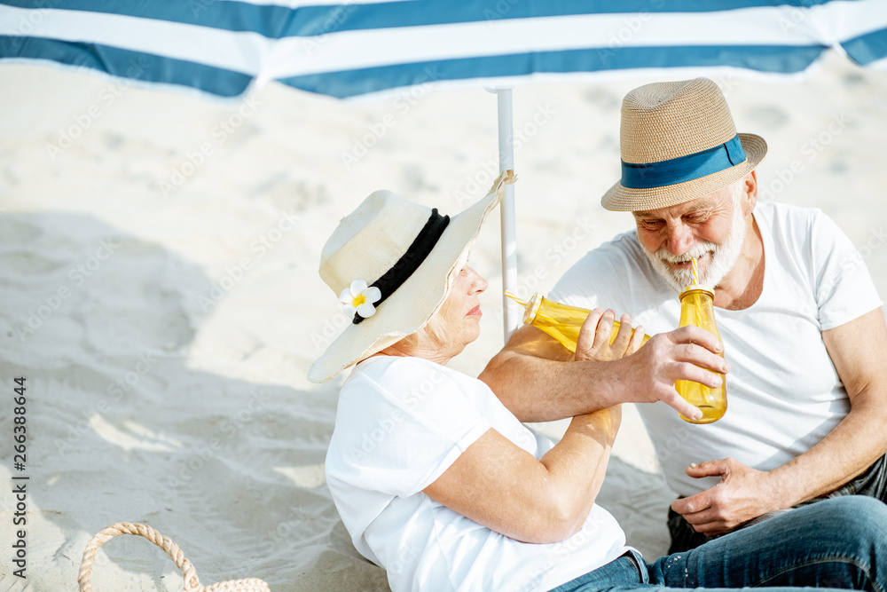 Happy senior couple relaxing, lying with drinks on the sandy beach, enjoying their retirement near t