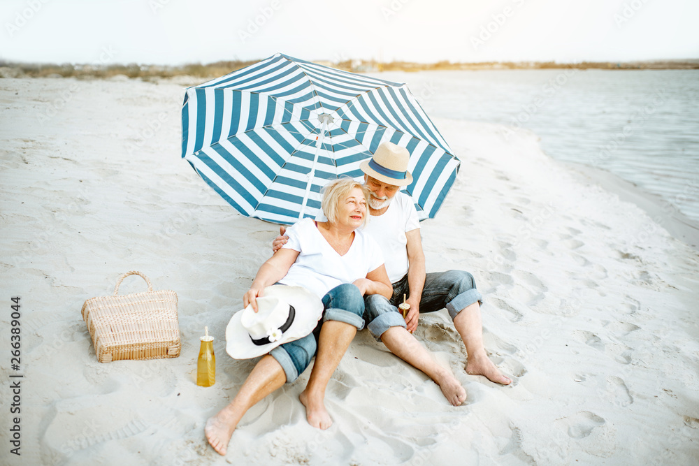 Happy senior couple relaxing, lying together under umbrella on the sandy beach, enjoying their retir