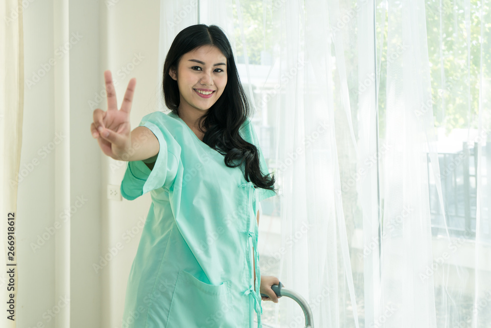 Young Asian patient woman standing at hospital room with walking stick showing victory sign for chee