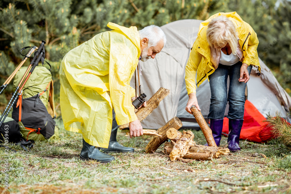 Senior couple in yellow raincoats making fireplace at the campsite near the tent in the woods