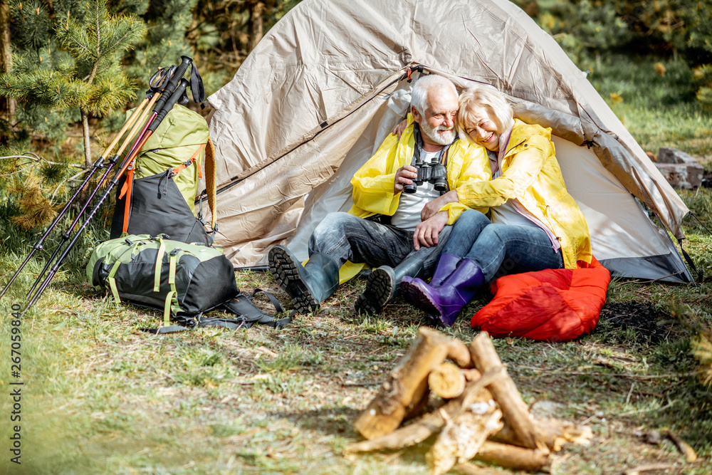 Senior couple in yellow raincoats sitting together near the tent at the campsite with fireplace in t