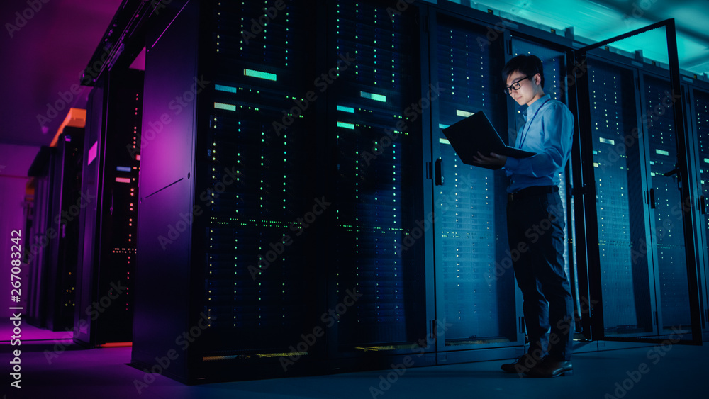 In Data Center: Male IT Technician Running Maintenance Programme on a Laptop, Controls Operational S