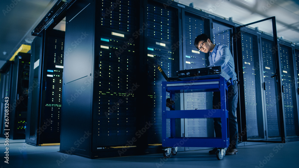 In Data Center: Male IT Technician Running Maintenance Programme on a Laptop, Controls Operational S