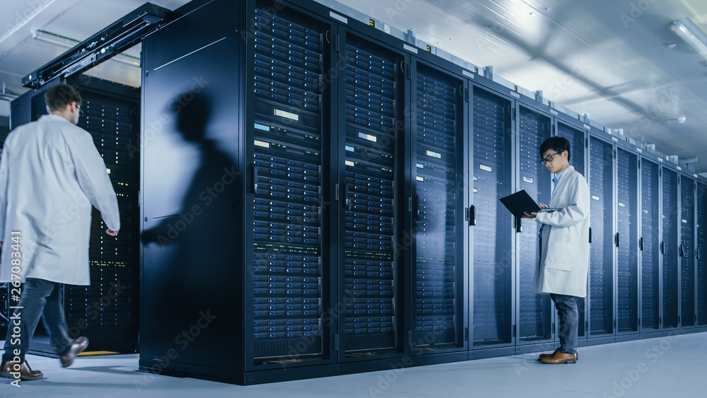 In Data Center: Male IT Technician Wearing White Coat Stands Beside Server Rack Cabinet, Uses Laptop