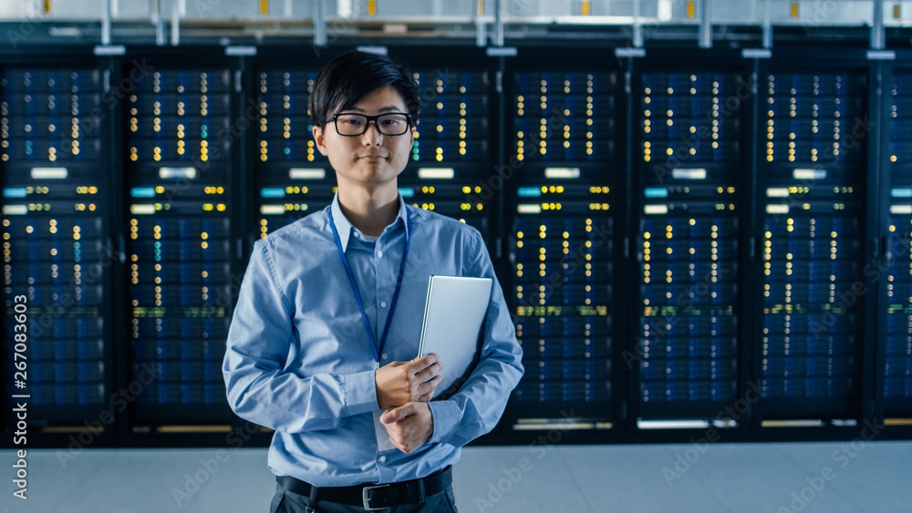 In the Modern Data Center: Portrait of IT Engineer Stands Beside the Row of Server Racks, Finishing 