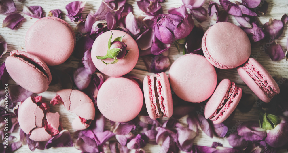 Flat-lay of sweet pink macaron cookies and rose buds and petals over wooden background, top view, cl