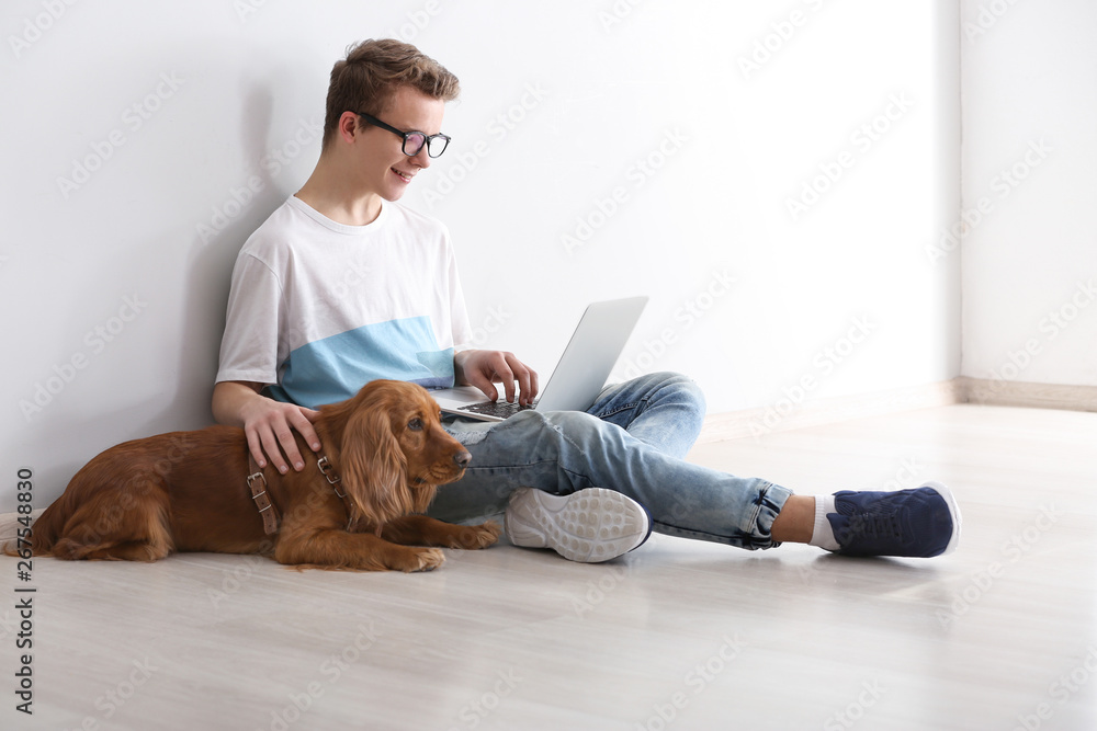 Teenage boy with cute dog and laptop sitting near light wall