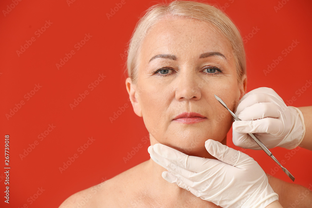 Mature woman and hands of plastic surgeon with lancet on color background