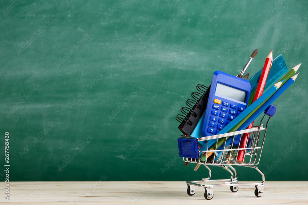 Education concept - school supplies in a shopping cart on the desk in the auditorium, blackboard bac