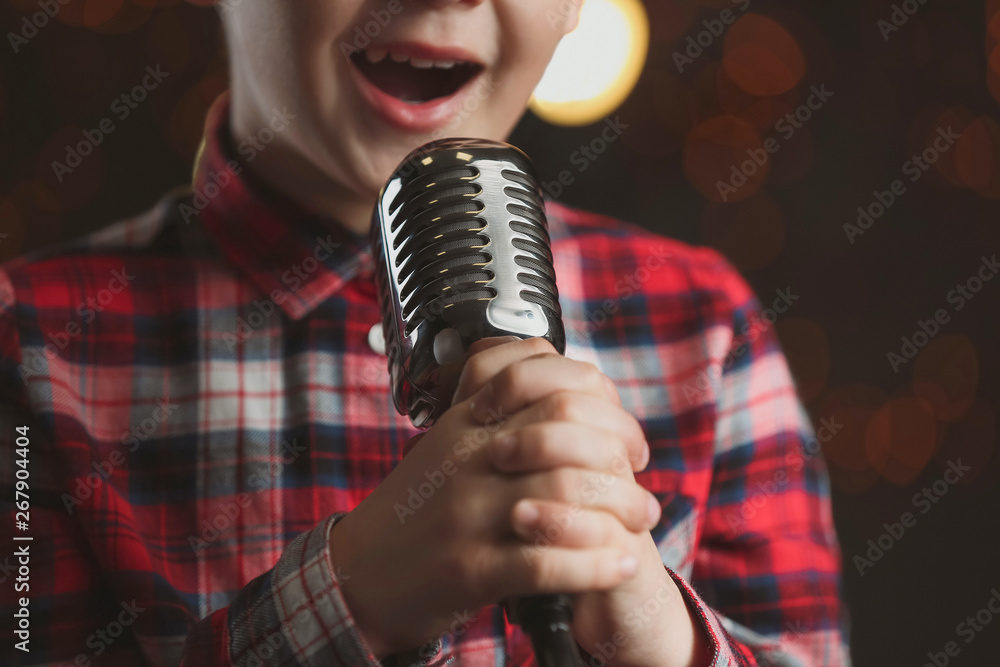 Cute little boy with microphone singing against dark background, closeup