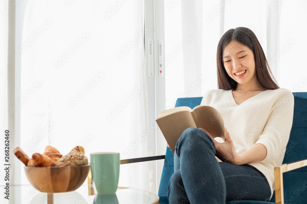 Young beautiful Asian woman relaxing in living room at home, reading books, drinking coffee in the m