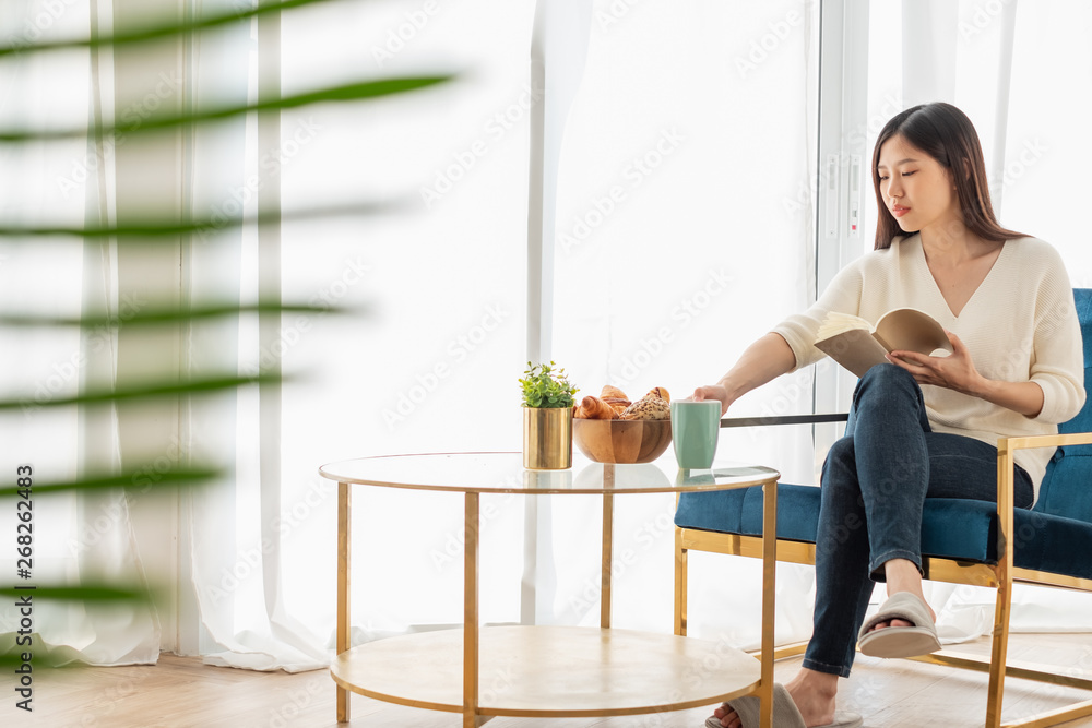 年轻漂亮的亚洲女人在家里的客厅里放松，看书，喝咖啡