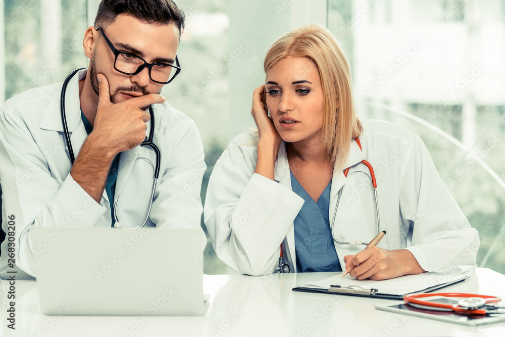 Young female doctor works at office in hospital while talking to male doctor sitting beside her. Med