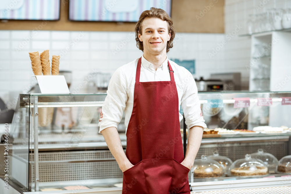 Portrait of a handsome salesman or waiter in red apron standing in the pastry cafe with shop-front o