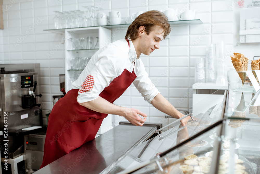 Young salesman in red apron placing sweet pastry in the refrigerator of the showcase at the modern c